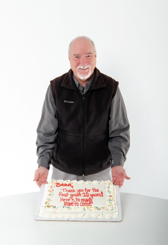 Sean smiling behind his anniversary sheet cake that reads, Sean, Thank you for the first great 10 years! Here's to many more to come.
