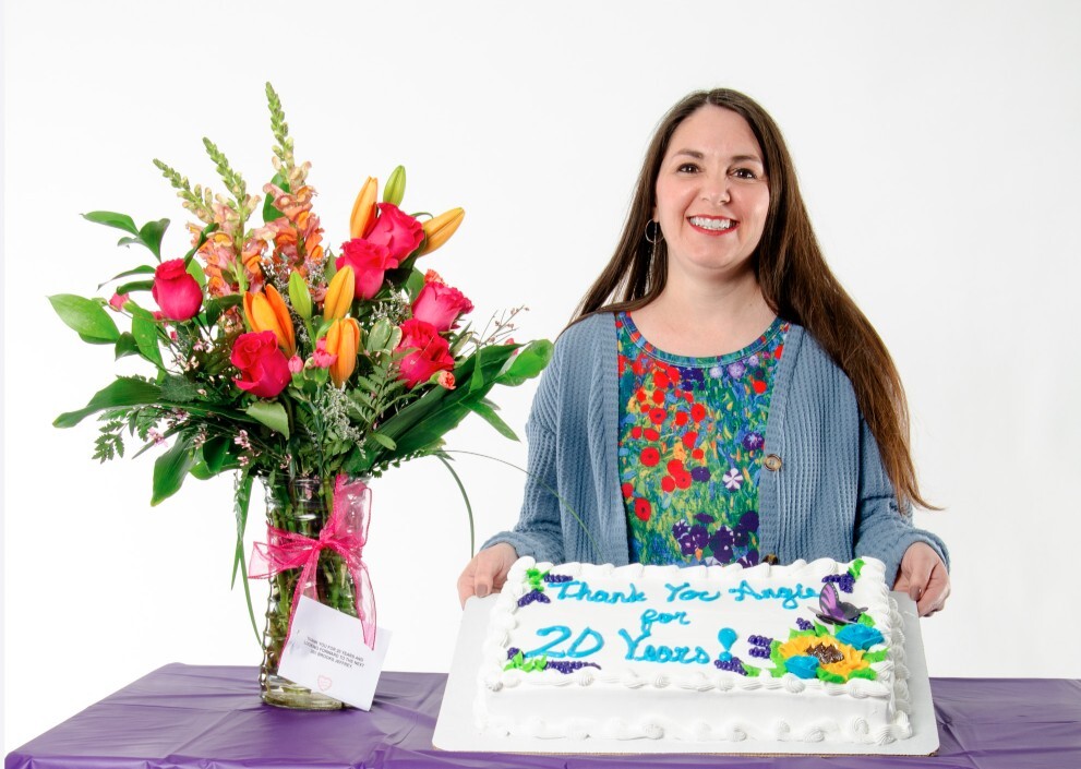 Angie holding a cake that says Thank you Angela for 20 years with a vase of colorful flowers next to her.